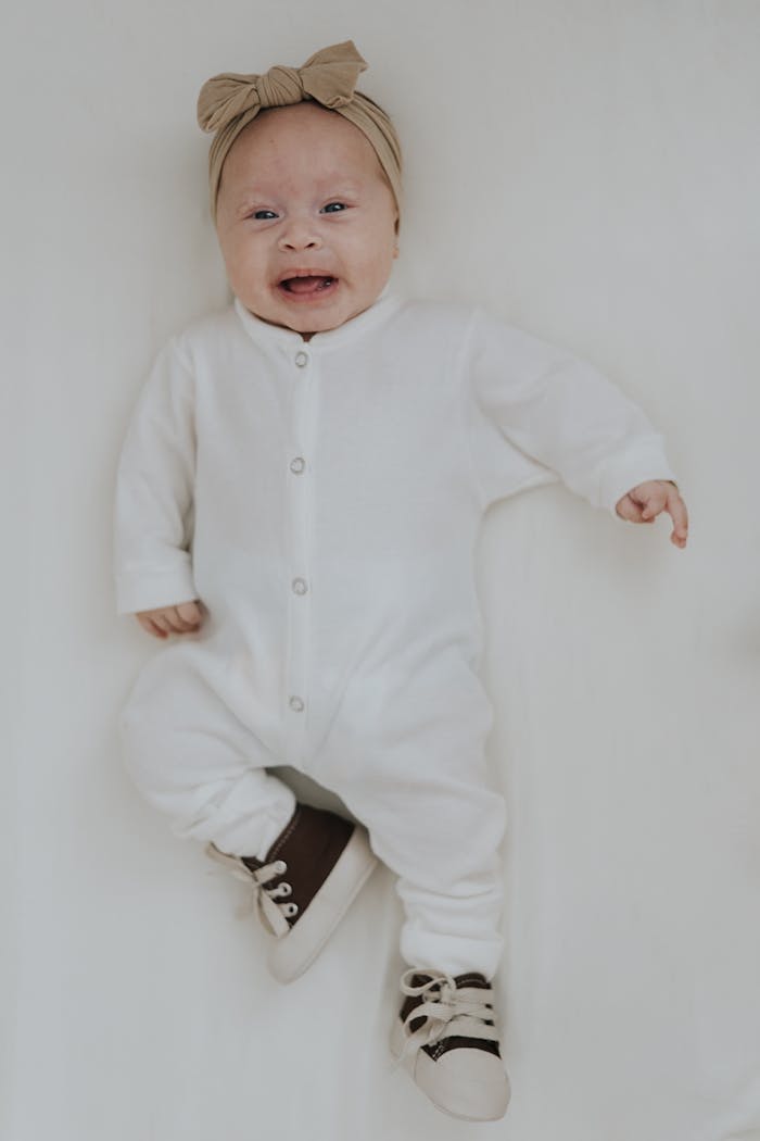 Cute baby in white bodysuit, wearing a bow headband and shoes, lying on a soft surface.